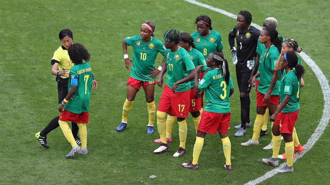 Mehrere Fußball-Nationalspielerinnen von Kamerun stehen zusammen mit einer Schiedsrichterin im Mittelkreises des Platzes beim WM-Spiel gegen England.