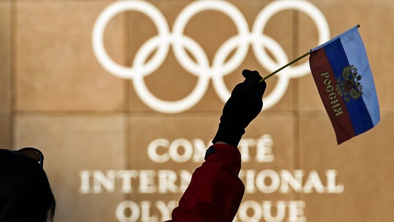 Eine Frau schwenkt eine russische Fahne vor den olympischen Ringen am Hauptquartier des Internationalen Olympischen Komitees (IOC) in Lausanne (Symbolbild). © picture alliance/Jean-Christophe Bott/KEYSTONE/dpa Foto: Jean-Christophe Bott