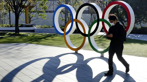 Eine Mann mit Mundschutz läuft an einem Aufsteller mit den Olympischen Ringen vorbei. © picture alliance / dpa 